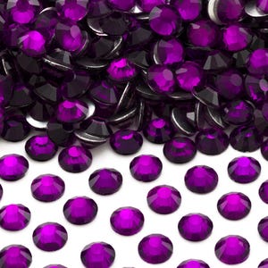 May include: A large pile of purple rhinestones, with a smaller group of rhinestones scattered on a white surface.
