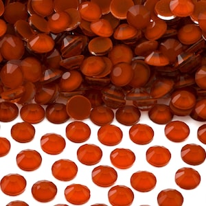 May include: A large pile of orange-colored rhinestones, with a smaller group of the same rhinestones spread out on a white surface.
