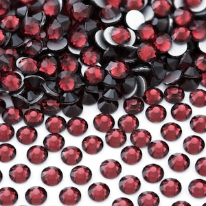 May include: A large quantity of small, round, red rhinestones. The rhinestones are scattered on a white surface.