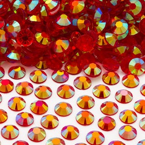 May include: A large pile of red iridescent rhinestones, with a smaller group of the same rhinestones spread out on a white surface.
