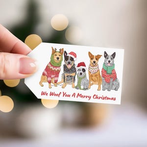 Peut inclure: Une étiquette cadeau rectangulaire blanche avec une illustration colorée de cinq chiens portant des pulls de Noël, des écharpes et des bonnets de Père Noël. Le texte "We Woof You A Merry Christmas" est imprimé en rouge sur le bas de l'étiquette.