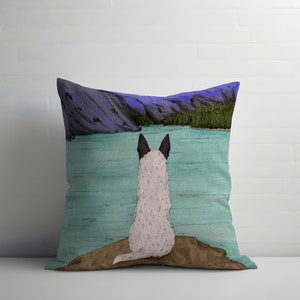 May include: A throw pillow with a blue and green watercolor design of a lake and mountains. A white and black dog sits on a rock in the foreground, looking out at the lake.