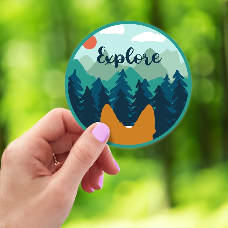 May include: Round sticker with a blue, green, and orange mountain scene. The text "Explore" is written in white on the sticker. The sticker is held in a hand.