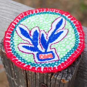 May include: A hand-stitched embroidery of a blue plant with a red and orange pot on a white background. The embroidery is surrounded by a red, green, and blue border.