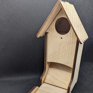 Op de afbeelding: Een houten dobbelsteentoren in de vorm van een huis met een scharnierend deksel en een lade om dobbelstenen te rollen. De toren is gemaakt van licht hout met een donkerbruine vlek rond de randen.