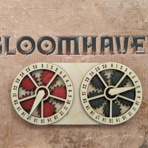 Puede incluir: Dos diales de madera en forma de engranaje con los números del 0 al 26. Los diales están conectados por una barra de madera. El texto "GLOOMHAVEN" está impreso sobre los diales.