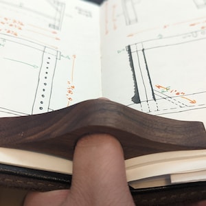 May include: A dark brown wooden page holder with a curved shape. The page holder is resting on an open notebook with handwritten drawings and notes.