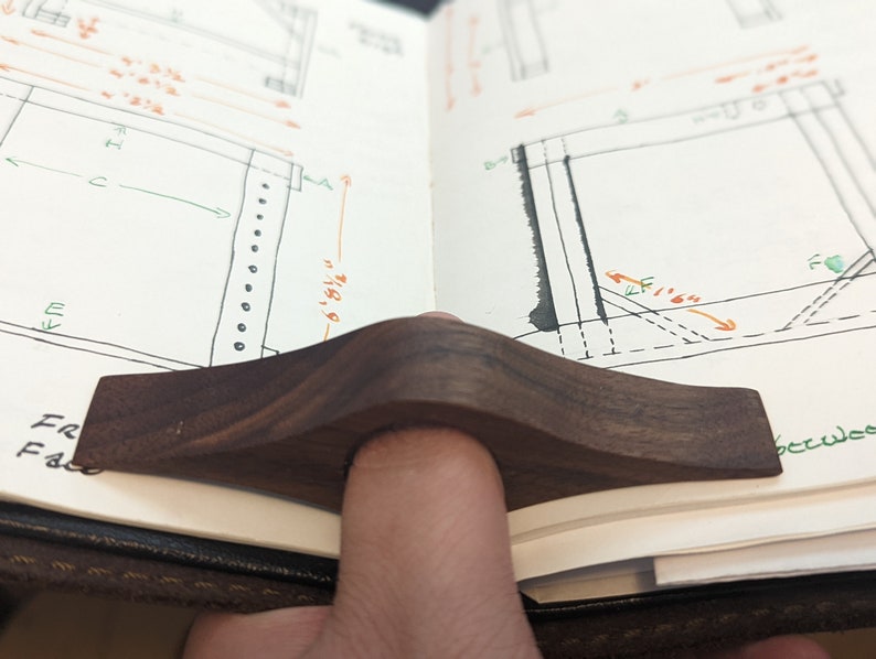May include: A dark brown wooden page holder with a curved shape. The page holder is resting on an open notebook with handwritten drawings and notes.
