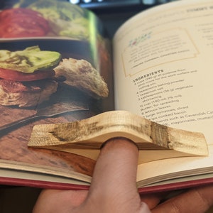May include: A close-up of a cookbook open to a recipe for turkey biscuits. The recipe includes ingredients such as all-purpose flour, baking powder, salt, shortening, milk, butter, lettuce, tomatoes, bacon, and Cavendish Catsup. A wooden finger ring is holding the page open.