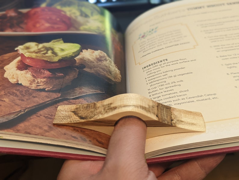 May include: A close-up of a cookbook open to a recipe for turkey biscuits. The recipe includes ingredients such as all-purpose flour, baking powder, salt, shortening, milk, butter, lettuce, tomatoes, bacon, and Cavendish Catsup. A wooden finger ring is holding the page open.