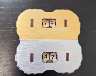 以下が含まれることがあります： 金色と銀色の仕上げが施された木製の数字カウンター2つ。上のカウンターは数字の7と9を表示し、下のカウンターは数字の4と7を表示しています。