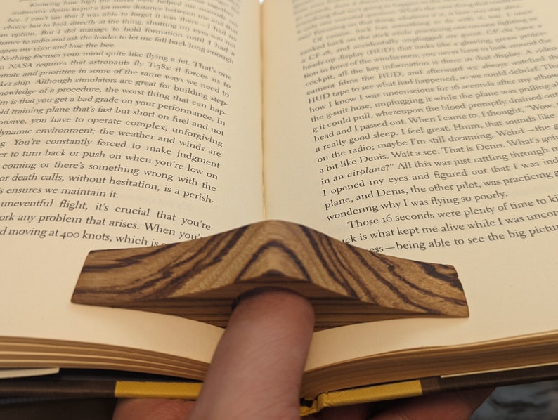 May include: A wooden book holder with a natural wood grain pattern. The holder is shaped like a wedge and is designed to hold a book open.