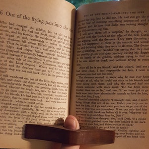 May include: A close-up of a book open to a page with the title "Out of the Frying-Pan into the Fire". The text describes a scene from the book, where a character is escaping from goblins. The book is held open by a wooden page holder.