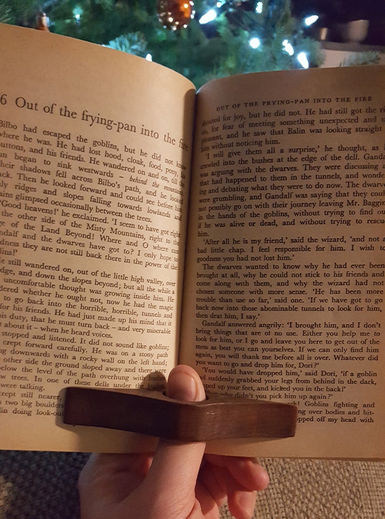 May include: A close-up of a book open to a page with the title "Out of the Frying-Pan into the Fire". The text describes a scene from the book, where a character is escaping from goblins. The book is held open by a wooden page holder.
