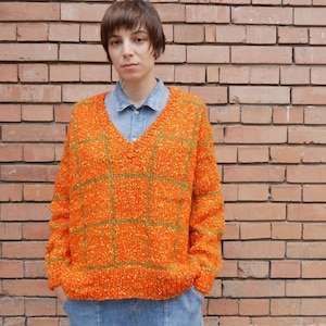 May include: A woman wearing a bright orange and green plaid jumper with a v-neck. The jumper has a loose fit and is made of a soft, textured material.