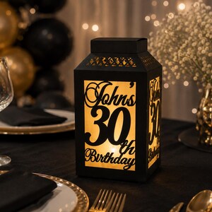 May include: A black rectangular lantern with a cutout design that glows from within. The front panel features the text John's 30th Birthday in a decorative script and bold font. The top section has a geometric pattern of small cutouts.