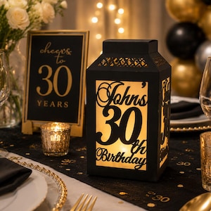 May include: A black rectangular lantern with cut-out lettering that reads John's 30th Birthday, illuminated from within by a warm light. The lantern features a decorative lattice pattern at the top and sits on a table decorated for a celebration.