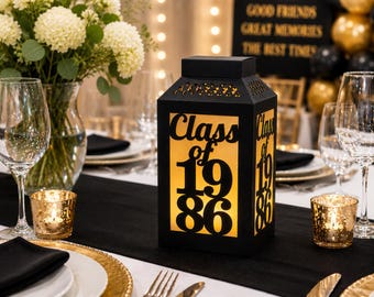 May include: A black rectangular lantern with a glowing interior featuring the cut-out text Class of 1986 on the front and side panels. The lantern has a square top and a decorative perforated border along the upper edge of the frame.