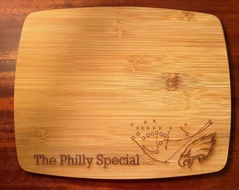 Puede incluir: Una tabla de cortar de madera con un acabado de grano de madera marrón. La tabla tiene un diagrama de juego de fútbol y un logotipo de águila grabados en ella. El texto "The Philly Special" también está grabado en la tabla.