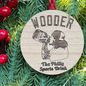 May include: A round wooden ornament with the text "WOODER" and "The Philly Sports Drink" printed on it. A cartoon image of a person drinking from a bottle labeled "WOODER" is in the center of the ornament.