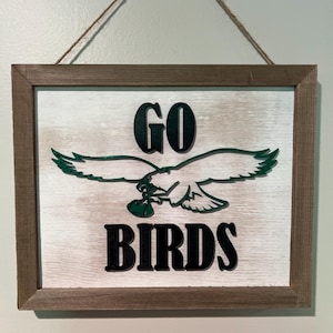 May include: A wooden sign with a white background and a brown frame. The sign says "GO BIRDS" in black letters with a green eagle holding a football in its talons.