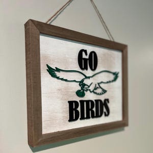May include: A wooden sign with a distressed white background and a brown frame. The sign features a green eagle with a football in its talons and the text "GO BIRDS" in black.