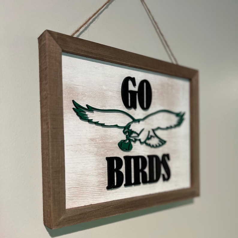 May include: A wooden sign with a distressed white background and a brown frame. The sign features a green eagle with a football in its talons and the text "GO BIRDS" in black.