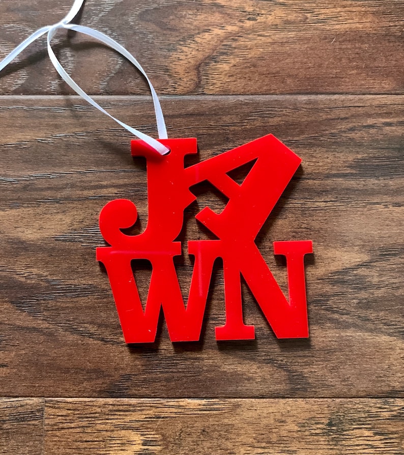 Op de afbeelding: Rode acryl ornament met het woord "JAWN" uitgesneden. De ornament hangt aan een wit lint.