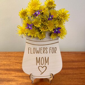 May include: A wooden sign with a vase shape and a bouquet of yellow dandelions and purple flowers. The sign says "Flowers for Mom" with a heart.