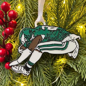May include: A decorative Christmas ornament featuring a football player in a green and white uniform, head down. The ornament is hanging from a white ribbon against a backdrop of evergreen branches and red berries.