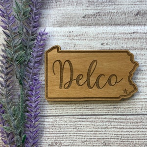 May include: Wooden sign in the shape of the state of Pennsylvania with the word "Delco" written in cursive script. A small star is in the lower right corner of the sign.