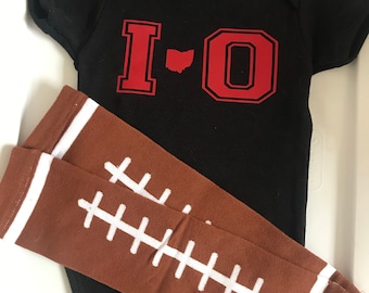 May include: A black baby onesie with the text "I-O" in red with an outline of the state of Ohio in between the letters. The onesie is paired with brown and white striped leg warmers that have a football stitch design.