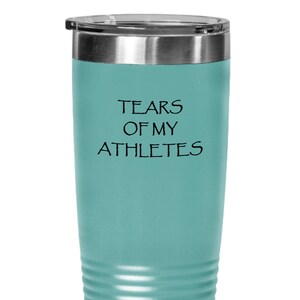 May include: A teal insulated tumbler with a silver rim and a black lid. The tumbler has the phrase "TEARS OF MY ATHLETES" printed in black. The tumbler is designed to keep beverages hot or cold.