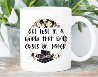 Puede incluir: Taza de cerámica blanca con asa negra. Presenta la cita "Get lost in a world that only exists on paper" dentro de una corona floral. También se muestra una pila de libros.