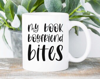 May include: White ceramic mug with the text "my book boyfriend bites" in black script. The mug has a curved handle and sits on a weathered wooden surface. Green leafy plants are in the background.