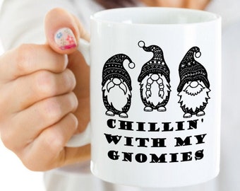 Könnte beinhalten: Weiße Keramik-Tasse mit drei Zwergen mit gemusterten Hüten und Bärten. Auf der Tasse steht in Schwarz der Text "CHILLIN' WITH MY GNOMIES". Die Tasse wird von einer Person gehalten.