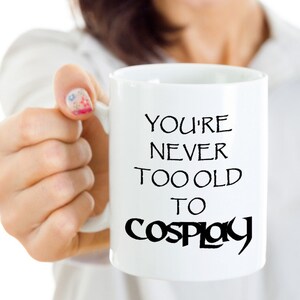 May include: White ceramic mug with black text that reads "You're never too old to cosplay".