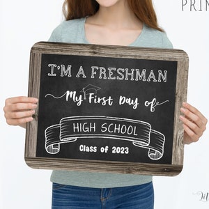 May include: A chalkboard sign with a wooden frame, held by a person. The sign reads "I'M A FRESHMAN My First Day of HIGH SCHOOL Class of 2023". The text "EDIT, DOWNLOAD AND PRINT NOW" is also visible.
