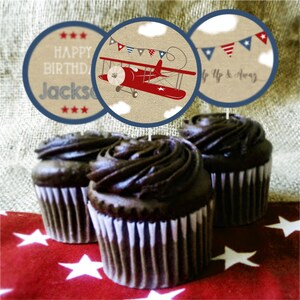 May include: Three chocolate cupcakes with brown frosting topped with round paper cupcake toppers. The toppers feature a red airplane with white clouds and blue stars. One topper says "Happy Birthday Jackson" and another says "Up Up & Away".