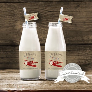 May include: Two glass milk bottles with labels that read "Up Up & Away" and "It's Jackson's 1st Birthday!" Each bottle has a red airplane graphic and a striped straw with a matching flag. The background is a wooden surface.
