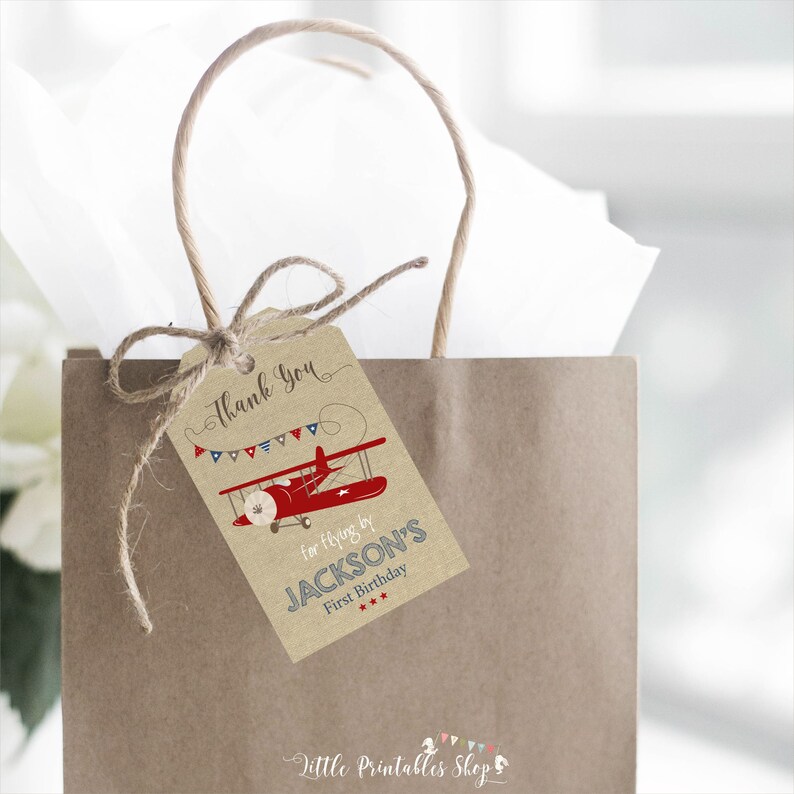 May include: A brown paper gift tag with a red aeroplane and blue and red bunting. The tag says "Thank You for flying by JACKSON'S First Birthday".