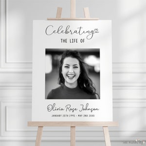 May include: A white sign on a wooden easel with the text "Celebrating THE LIFE OF" above a black and white photo of a smiling woman. Below the photo is the name "Olivia Rose Johnson" and dates.