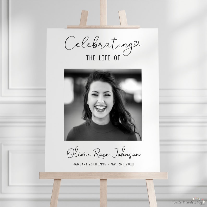 May include: A white sign on a wooden easel with the text "Celebrating THE LIFE OF" above a black and white photo of a smiling woman. Below the photo is the name "Olivia Rose Johnson" and dates.