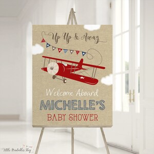 May include: A burlap welcome sign with a red airplane and blue and red bunting. The sign reads "Up Up & Away" and "Welcome Aboard Michelle's Baby Shower".