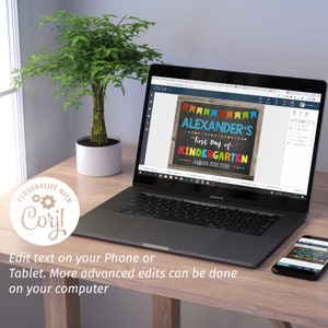 May include: A laptop displays a colorful chalkboard-style sign that reads "ALEXANDER'S First Day of KINDERGARTEN August 25th 2019." The sign is framed with a wooden border and decorative pennants. A smartphone shows the same design. A small potted plant sits nearby.