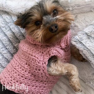 May include: A small, brown and black dog wearing a pink crocheted sweater. The dog is lying on a white and gray fuzzy surface. The sweater has a hole for the dog's head and two holes for the dog's front legs. The sweater is crocheted in a simple stitch. The dog is looking at the camera. The text "Hookfully" is visible in the bottom left corner of the image.