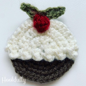 May include: A crocheted Christmas pudding ornament with white icing, brown cake, and a red berry with green leaves on top.
