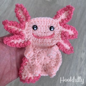 May include: A pink crocheted axolotl with black eyes and a pink and white crocheted tail. The axolotl is smiling.