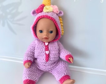 May include: A purple crocheted unicorn costume for a doll. The costume features a pink hood with a yellow horn and a pink and yellow mane. The costume has pink trim and white heart-shaped buttons.