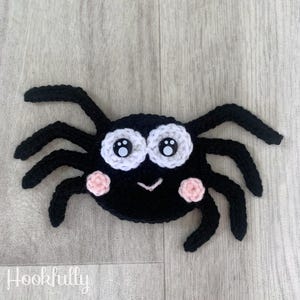 May include: A black crocheted spider with white eyes and pink cheeks. The spider has eight legs and a smiling face.